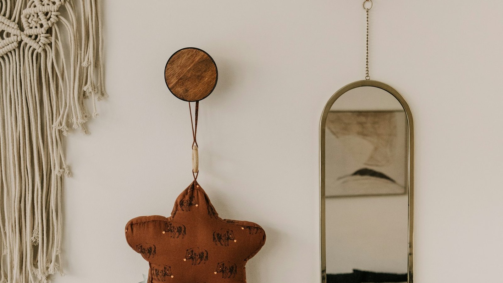 A nursery wall in neutral tones with a macrame hanging, a soft terracotta star cushion and a brass mirror