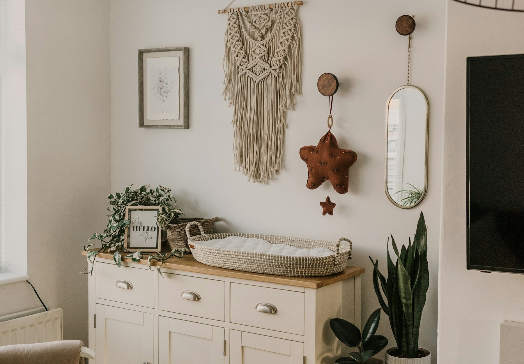 A calm nursery in cream and natural wood: a changing table, a hanging macrame, a woven basket and potted plants